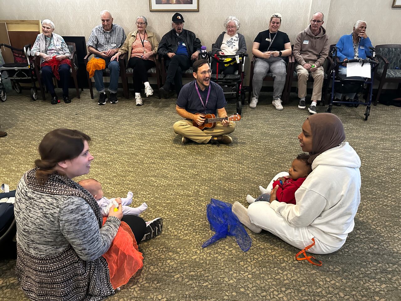 Seniors are seated in the background, a librarian seated on the ground sings with a ukelele and caregivers with children singalong.