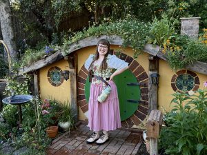 picture of a smiling woman standing in front of a low, yellow building with a green door. Plants cover the roof and ground surrounding the building.