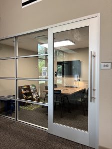 picture of a room with glass wall and door. Inside the room is a table with 5 chairs, a TV, a small table, a bookcase with toys and books, and a play rug.
