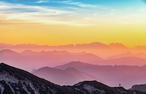 Photograph of a horizon with shades of pink, yellow, and blue. Mountains in the background with a hiker visible.
