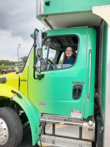 A middle age woman sitting in the driver seat of a Bookmobile cab. The Bookmobile is yellow and green.