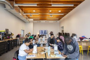 A bright makerspace or creative workshop with high ceilings, exposed wood beams, and large worktables. People of various ages are seated and standing around sewing machines, fabric, and craft materials, working on hands-on projects. Tool storage cabinets and equipment line one side of the room, while shelves and workstations fill the space. The environment is collaborative and educational, with participants engaged in learning, creating, and making together.