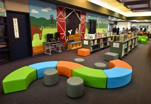 Elementary school library with colorful, modular seating arranged in a circular pattern in the foreground. Bookshelves filled with children’s books line the center of the room, while a large farm-themed mural featuring a barn, horse, windmill, and green fields decorates the wall. A computer workstation sits beneath the mural, and several people are visible in the background near reading and activity areas. The space is bright, open, and designed to encourage reading, collaboration, and learning.