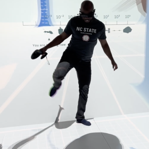 A person wearing a virtual reality (VR) headset and holding a handheld controller balances on one leg while interacting with an immersive virtual environment projected around them. They are wearing an NC State College of Engineering T-shirt, and the digital display behind them features graphics and a scale, suggesting an interactive educational or scientific VR experience.