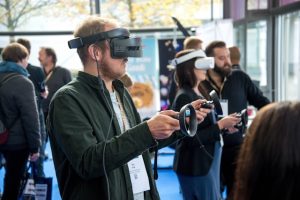 People wearing virtual reality (VR) headsets use handheld controllers to experience immersive technology during a technology expo or demonstration, while other attendees converse in the background.