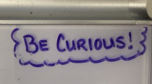 A white board with the phrase "Be curious!" written in purple ink.