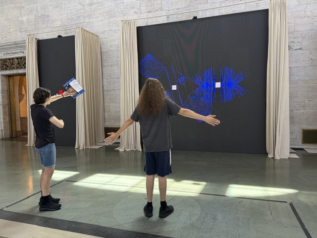 Two museum visitors stand in front of a large interactive digital art installation at the Detroit Institute of Arts. One visitor faces the screen with their arms outstretched while the other raises a brochure toward the display. The screen shows bright blue abstract light patterns that respond to their movements in a spacious gallery with stone walls.