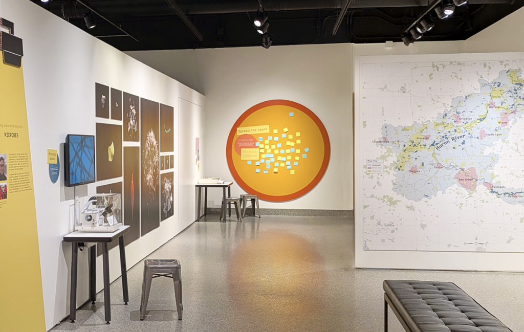 Interior view of a museum gallery featuring interactive exhibits. A large circular orange display on the back wall invites visitors to post sticky-note responses, while a large regional map, scientific images, display tables, stools, and benches encourage exploration and participation throughout the space.