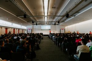 A large audience sits in rows facing a speaker presenting on stage in a modern conference hall. A presentation slide is projected on a large screen at the front of the room, with attendees listening to the talk in a professional learning environment.