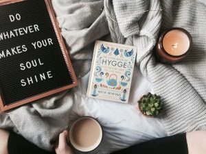 An overhead view of a cozy reading setup on a bed with soft gray knit blankets. A black letter board reads, "DO WHATEVER MAKES YOUR SOUL SHINE." Beside it is a copy of The Little Book of Hygge by Meik Wiking, a lit candle, a small potted succulent, and a mug of coffee held by a person sitting under the blankets. Warm string lights are draped across the scene, creating a calm, inviting atmosphere.