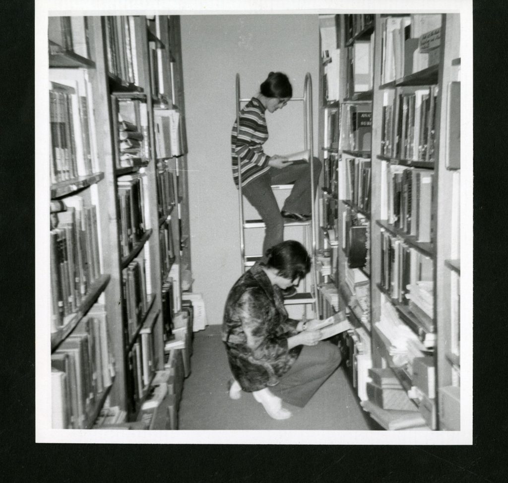 Two students reading in a library stack. One is elevated on a step stool. Photograph is in black and white