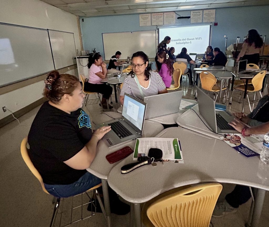 Women sit in desks with laptops in front of them within a classroom 