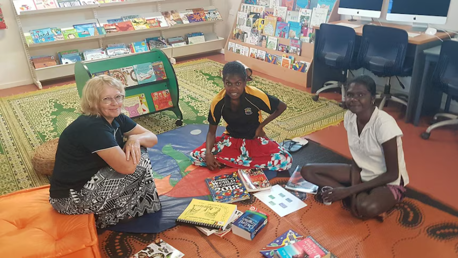 Three people sit on a library floor with a pile of books between them.