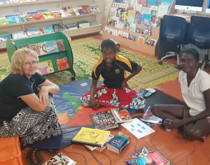 Three people sit on a library floor with a pile of books between them.