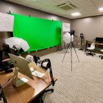 A green screen, camera, and lights set up in a room with computer stations.
