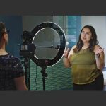 A woman stands in front of a camera and light to record a video.