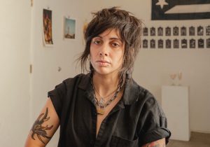Jo Cosme, one of the 2026 Artists in Residence at Seattle Public Library, is pictured here in a headshot with a black top and artwork in the backround.