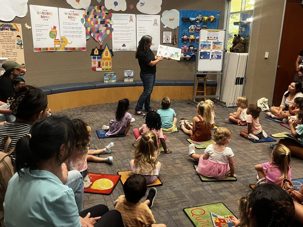 the mayor of a southern California city stands at the front of a room, reading a storybook to a group of kids at the library