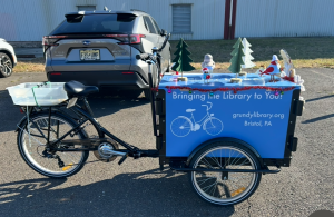 A three-wheeled bicycle with a blue box affixed to the front. The box is decorated for the holidays with small birds and trees. 