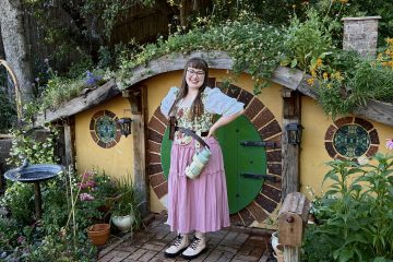 picture of a smiling woman standing in front of a low, yellow building with a green door. Plants cover the roof and ground surrounding the building.