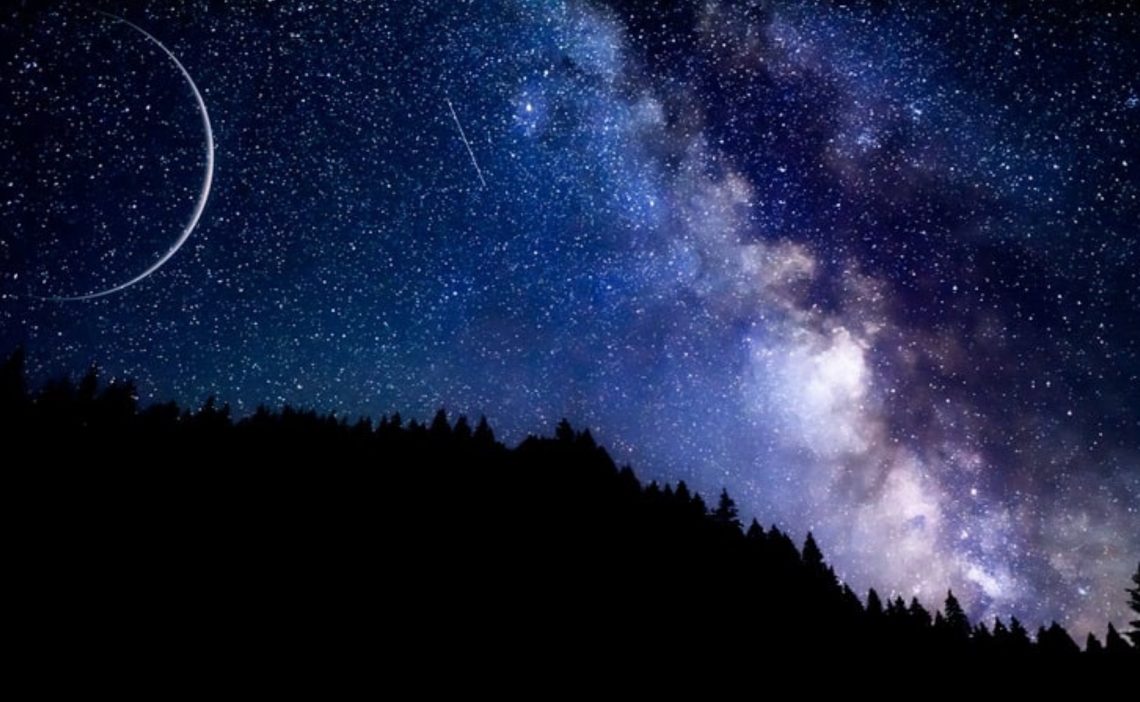 View of stars and clouds and a sliver of the moon behind a forest silhouette.