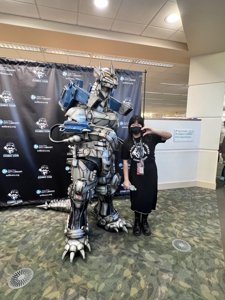 A picture of me at our library's Comic Con. I am standing next to a Godzilla mecha holding up a peace sign.