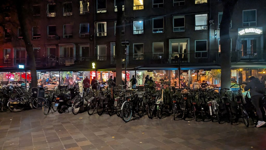 Shows a crowded bike parking space, with busy urban restaurants nearby.
